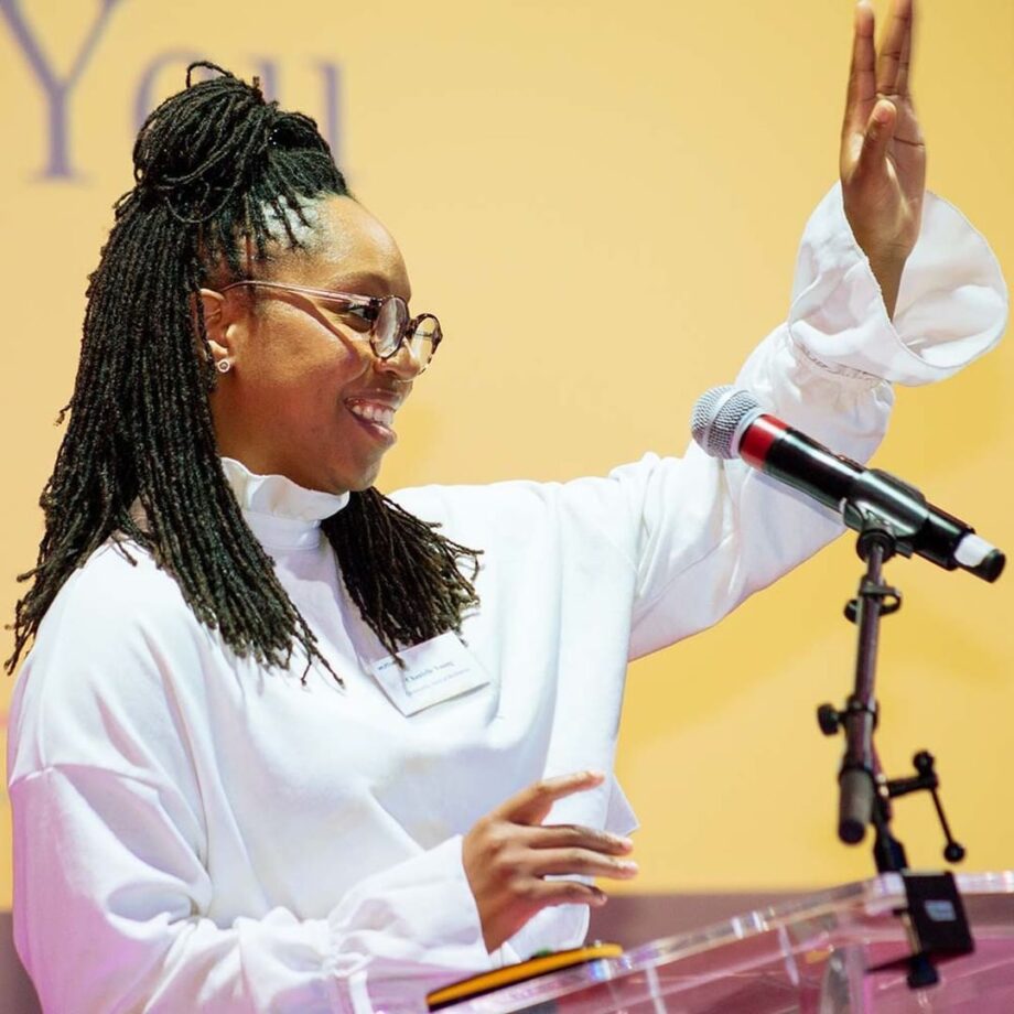 A young woman in a white shirt and glasses speaking into a microphone and waving